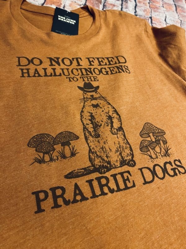 Close up view of the do not feed hallucinogens text above a prairie dog with a cowboy hat surrounded by mushrooms with prairie dogs text below t-shirt design